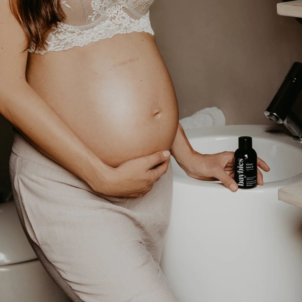 Schwangere Frau hält eine kleine Flasche Baybies Schwangerschafts Öl neben einem Waschbecken.