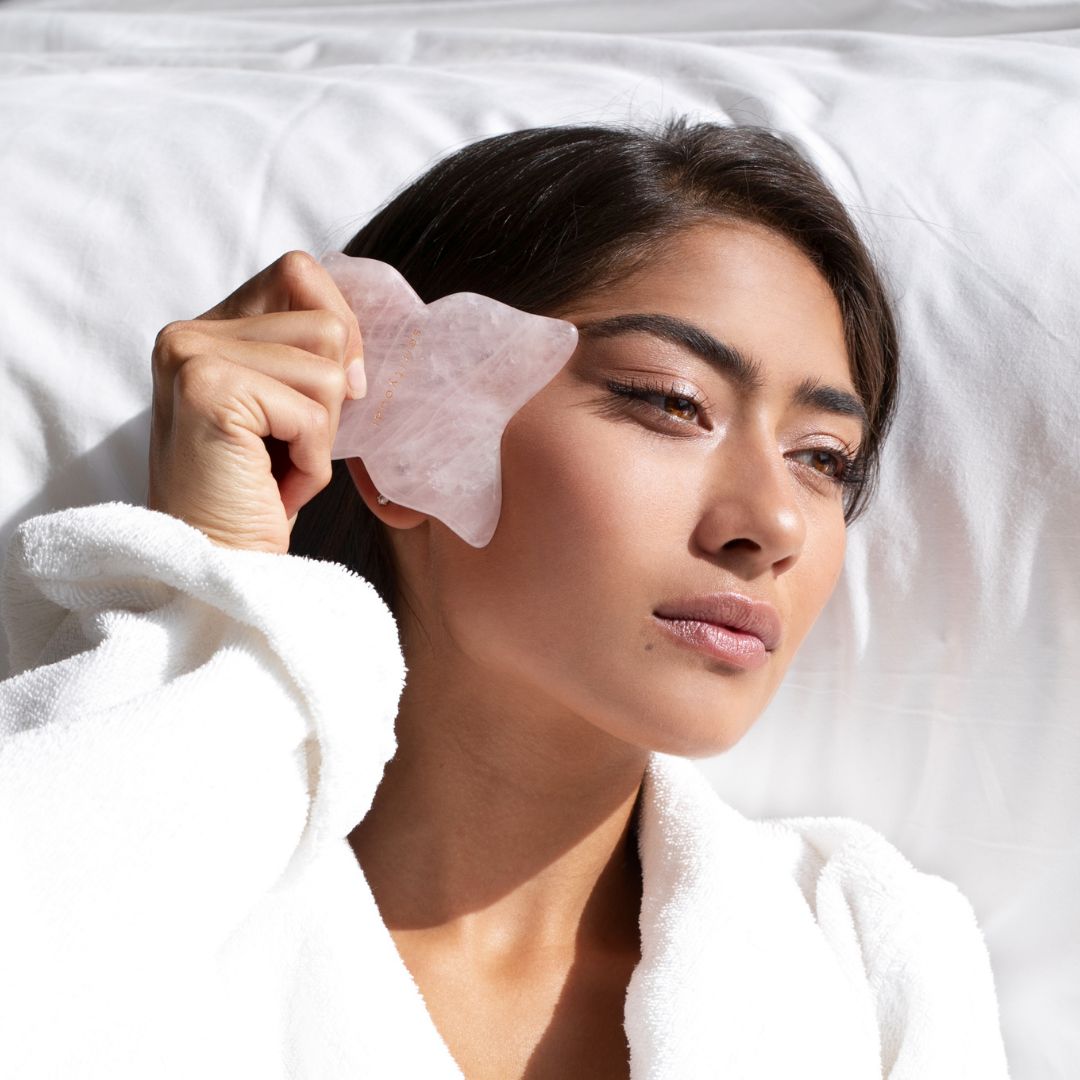 Frau mit rosa Gua Sha in Schmetterlingsform. Liegt auf weißer Bettwäsche.