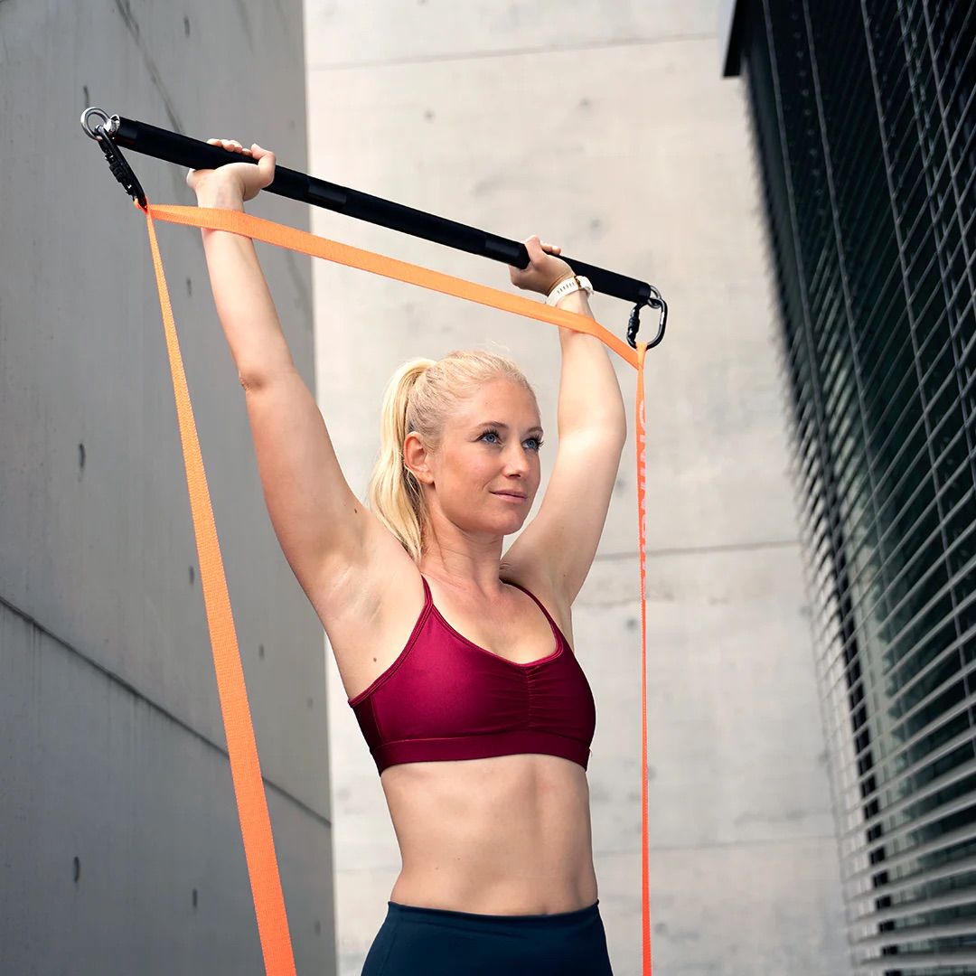 Frau mit Fitnessband und Stange. Sie steht vor einer Betonwand und hebt die Arme. Orangefarbenes Band.