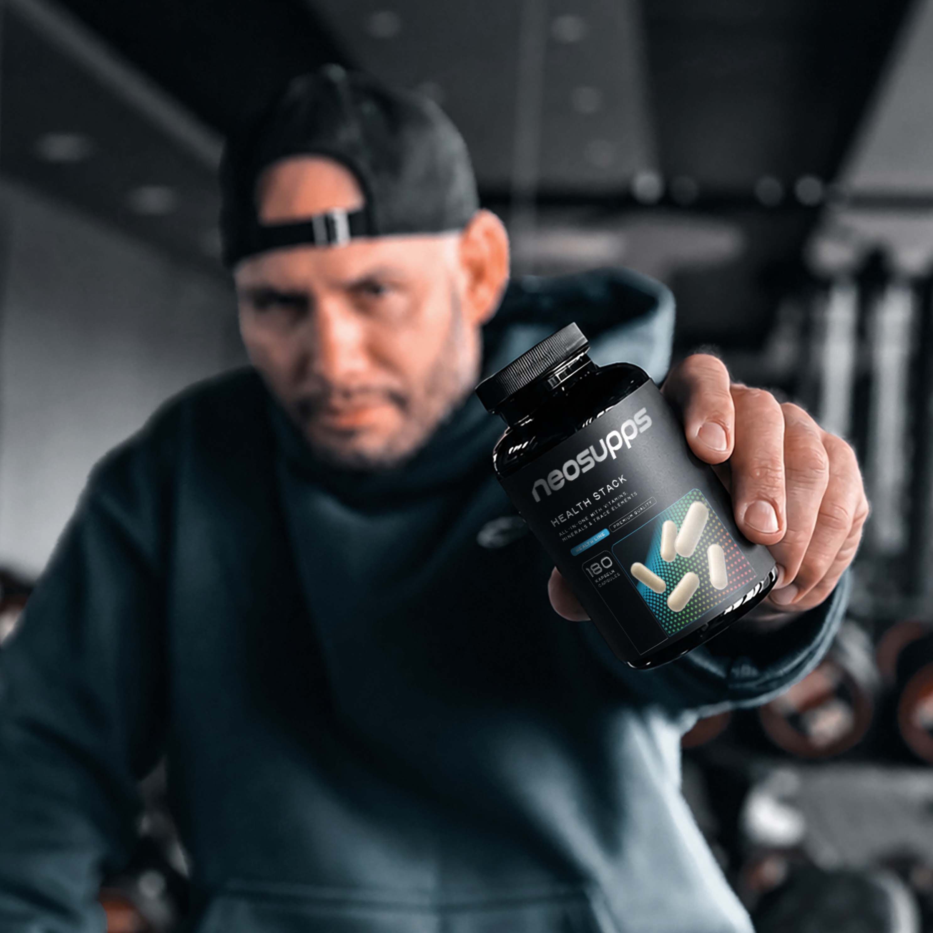 Mann hält Neosupps Health Stack Dose in der Hand, Fitnessstudio-Hintergrund.