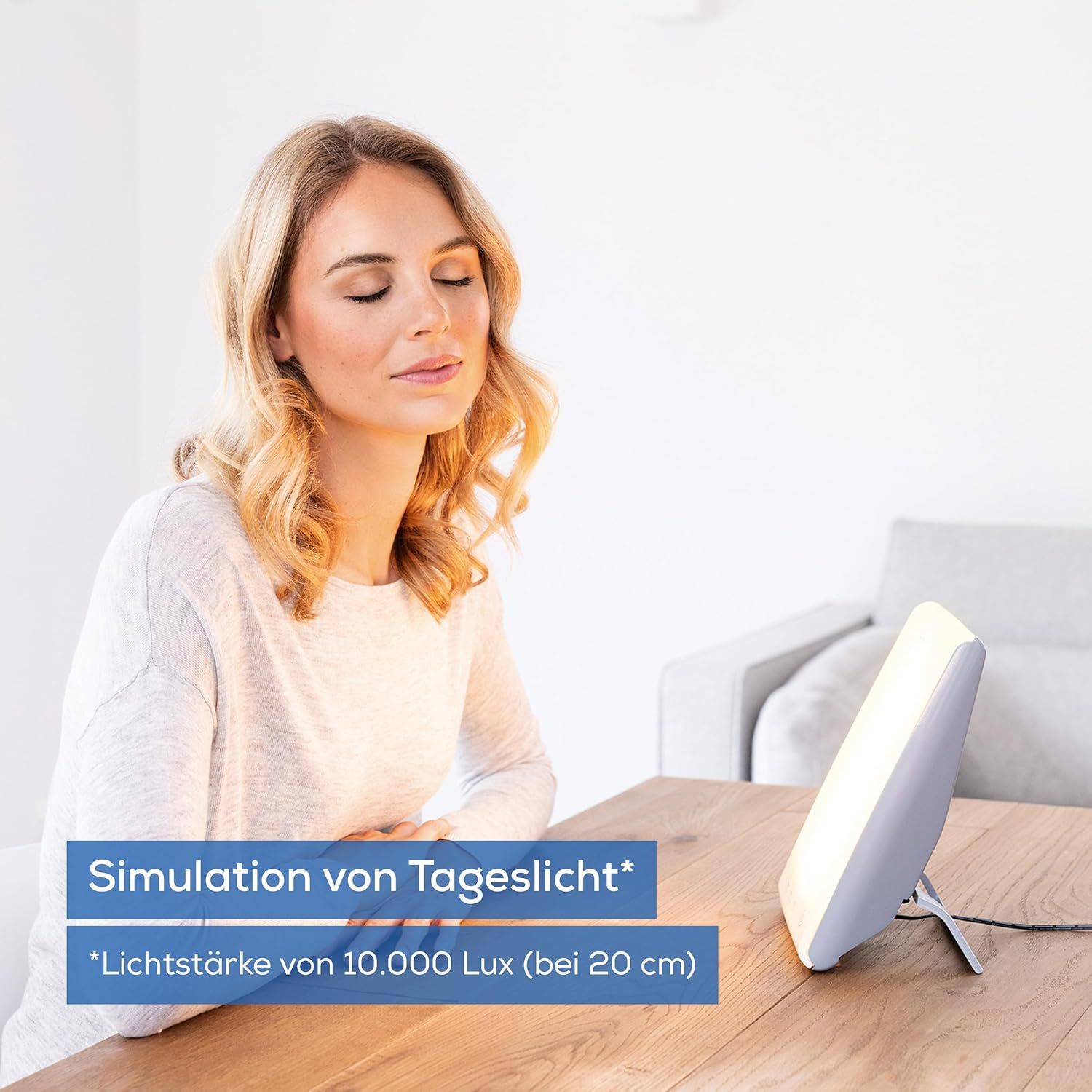 Tageslichtlampe auf einem Tisch. Eine Frau sitzt daneben. Text: Simulation von Tageslicht, Lichtstärke von 10.000 Lux.