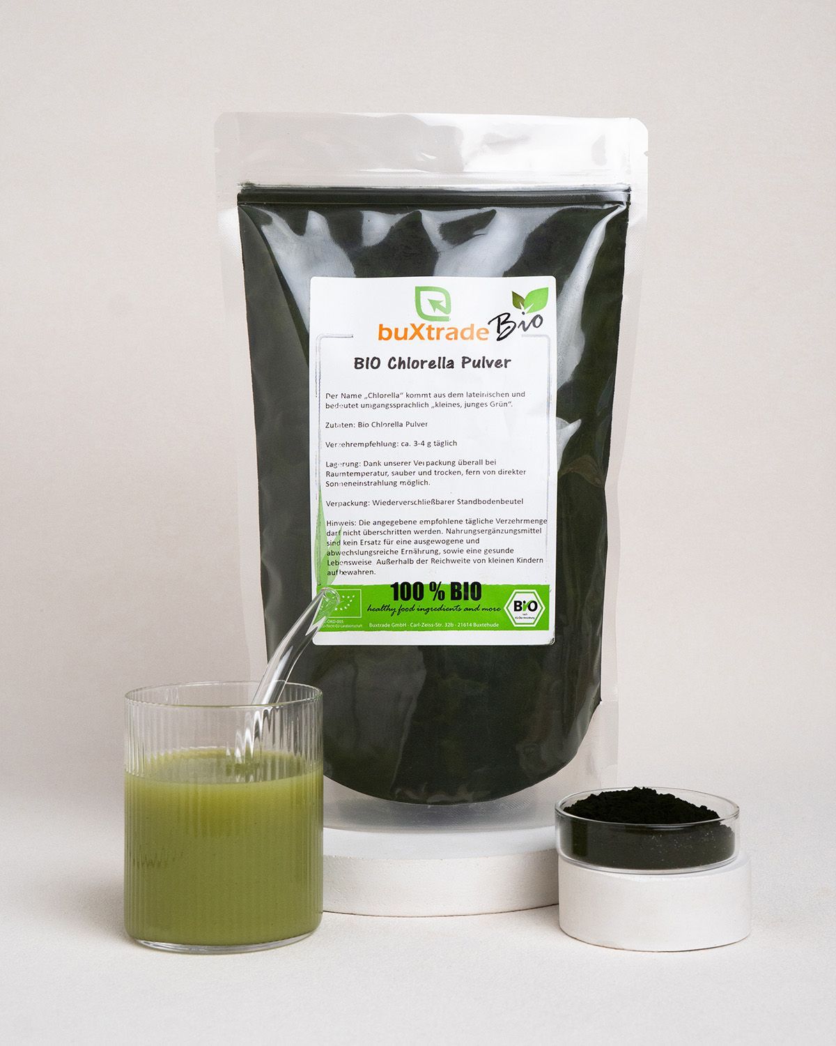 Glas mit grünem Getränk, daneben offenes Gefäß mit grünem Pulver und Verpackung von buXtrade Bio Chlorella Pulver.