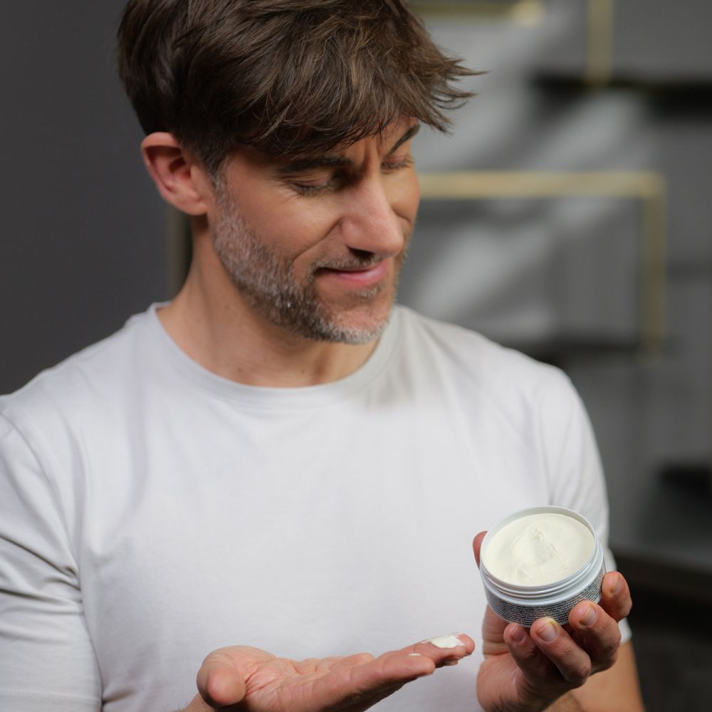 Mann hält Genki Styling Paste Dose. Grauer Deckel, schwarzer Schriftzug, gelber Farbfleck. Creme.