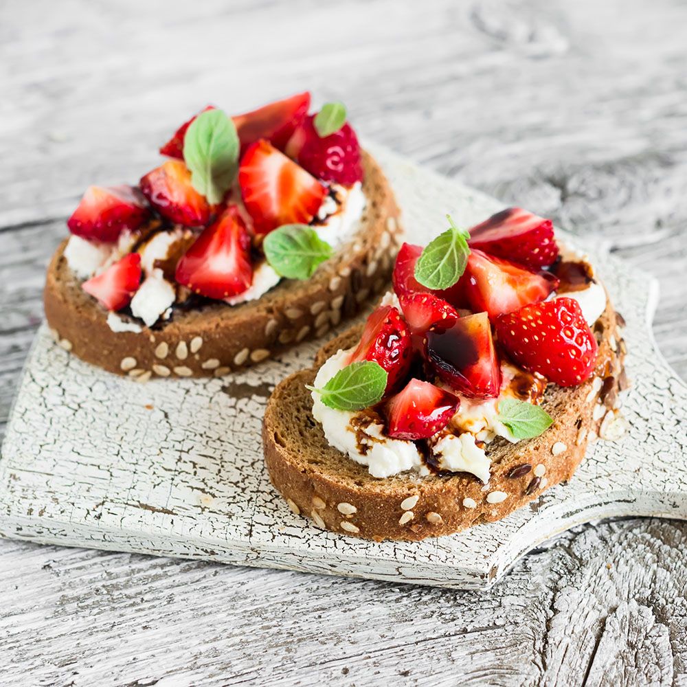 Zwei Brotscheiben mit Erdbeeren, Frischkäse und Balsamico-Glasur. Dekoriert mit Minze auf einem Holzbrett.