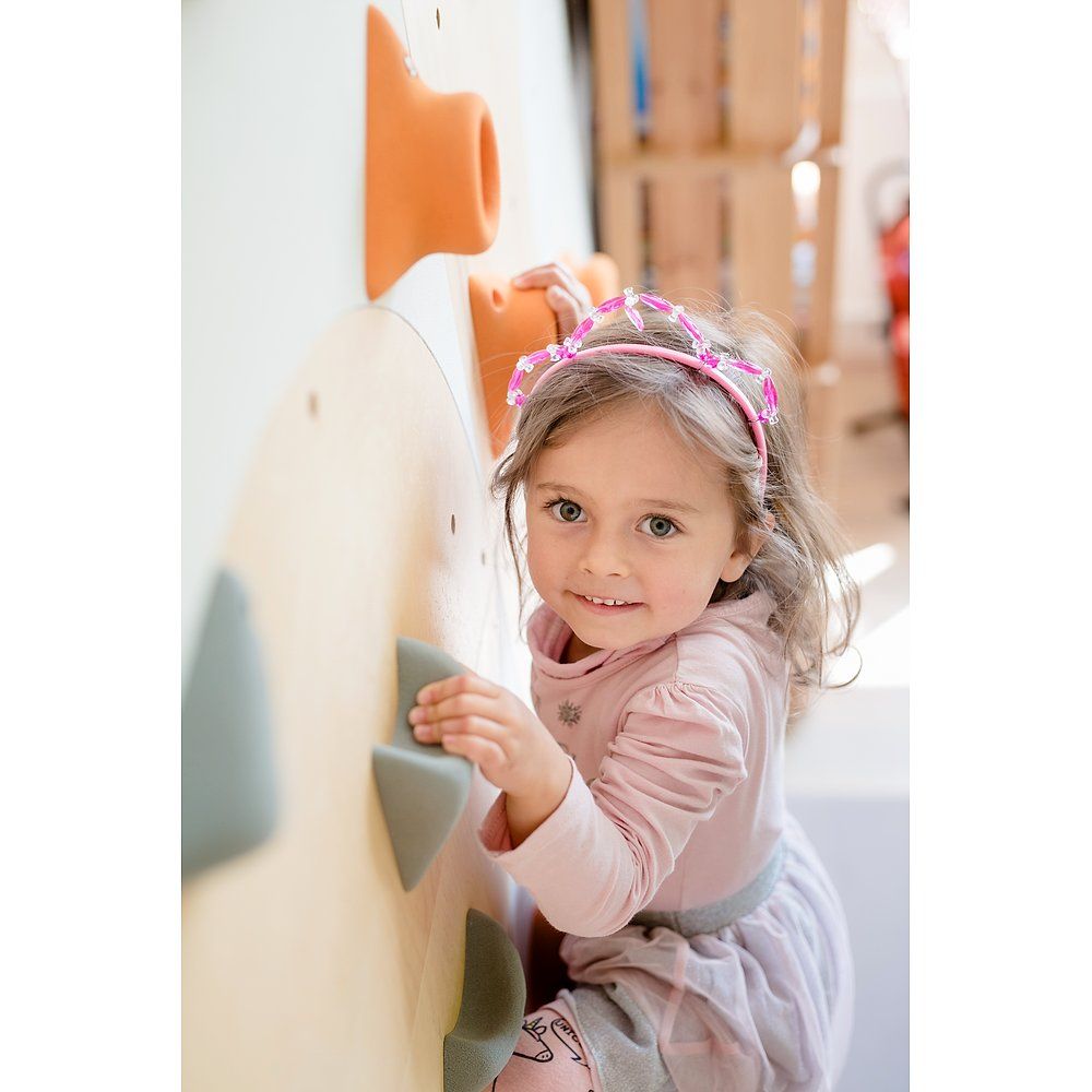 Mädchen klettert an einer Kletterwand mit bunten Griffen. Sie trägt ein Stirnband und ein rosa Kleid.