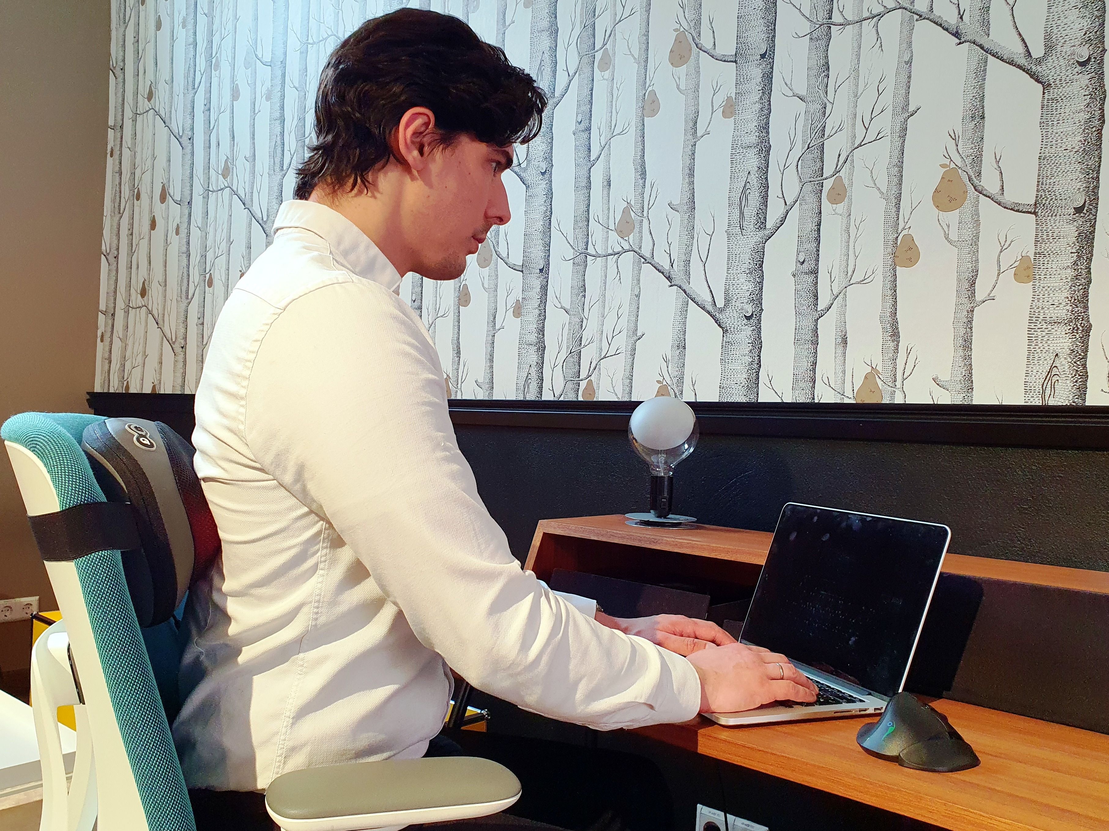 Mann sitzt an Schreibtisch, tippt auf Laptop. Ergonomischer Stuhl. Hintergrund: Tapete mit Baumdesign, Schreibtisch.
