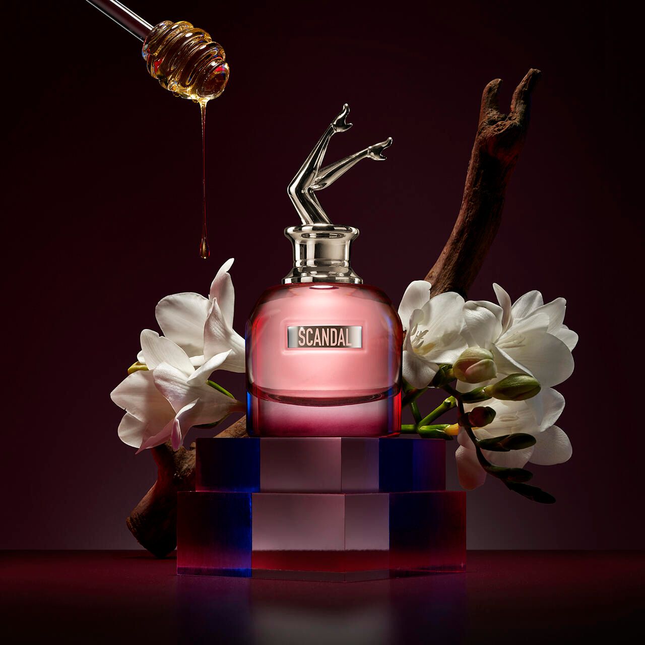 Pinkfarbener Flakon mit silbernem Verschluss und Bein-Anhänger. Aufschrift: SCANDAL. Umgeben von Blumen, Holz und Honig.