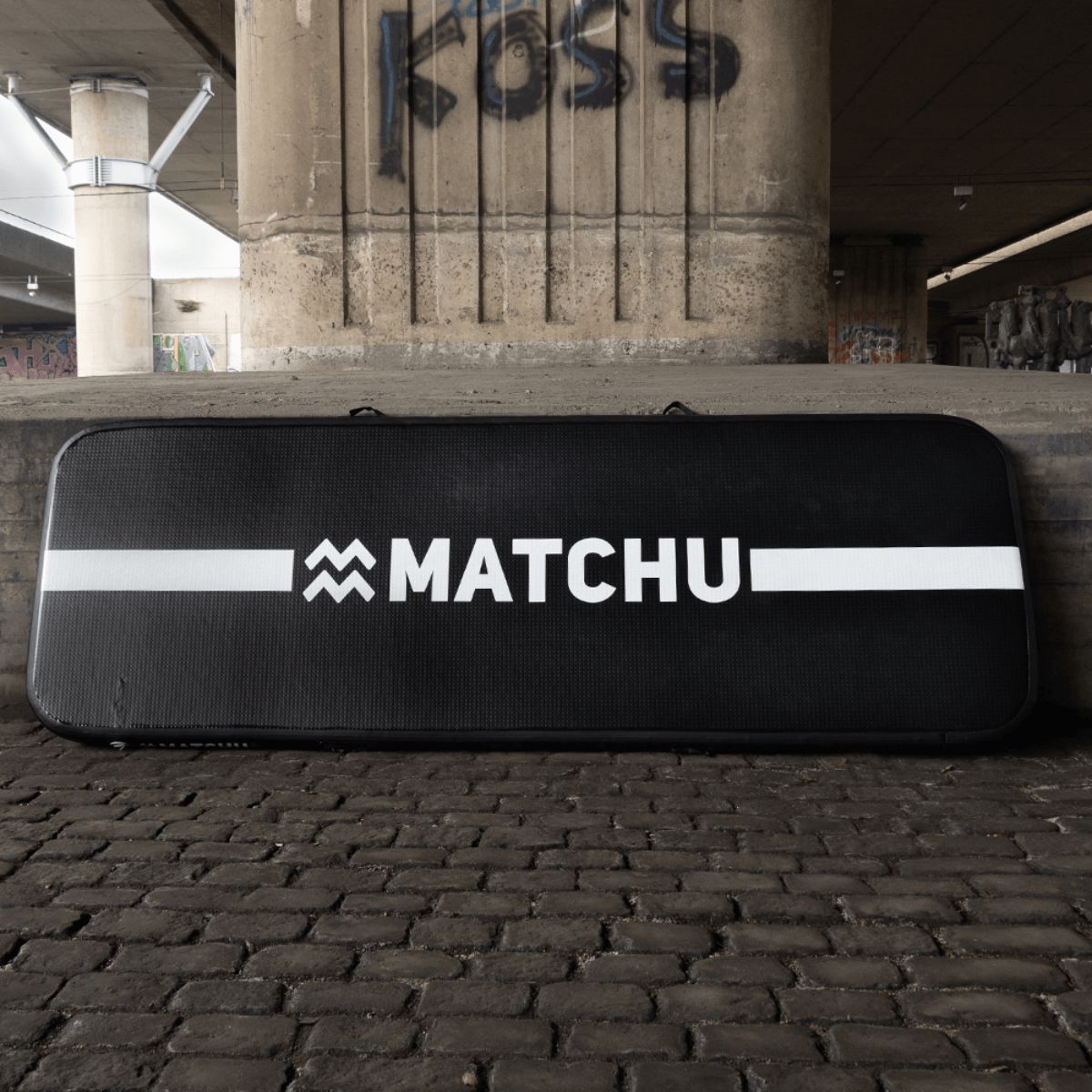 Schwarze Gymnastikmatte mit weißem Streifen und Logo. Marke: MATCHU. Liegt auf Pflastersteinen.