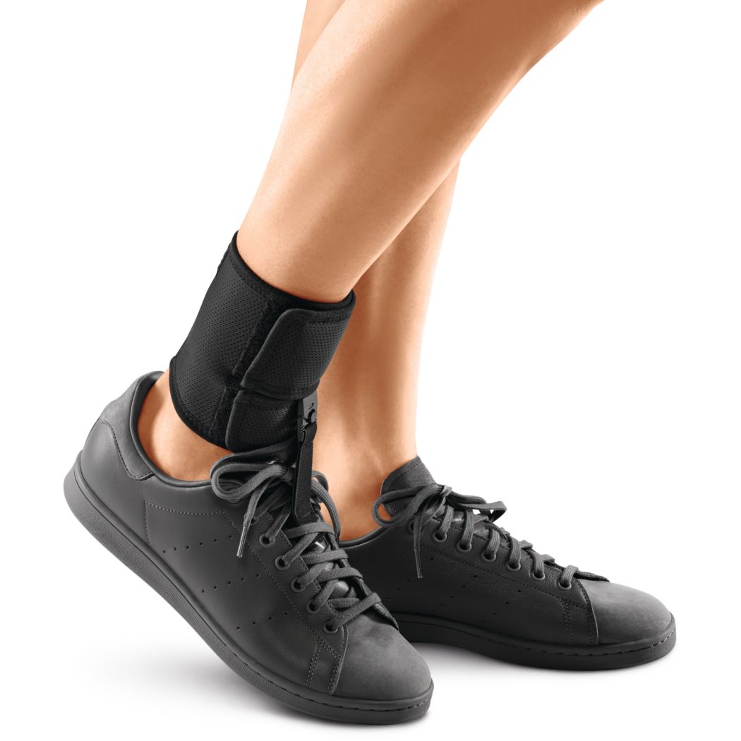 Fußheberorthese am Knöchel einer Person. Schwarz, mit Schuh. Füße in schwarzen Turnschuhen.