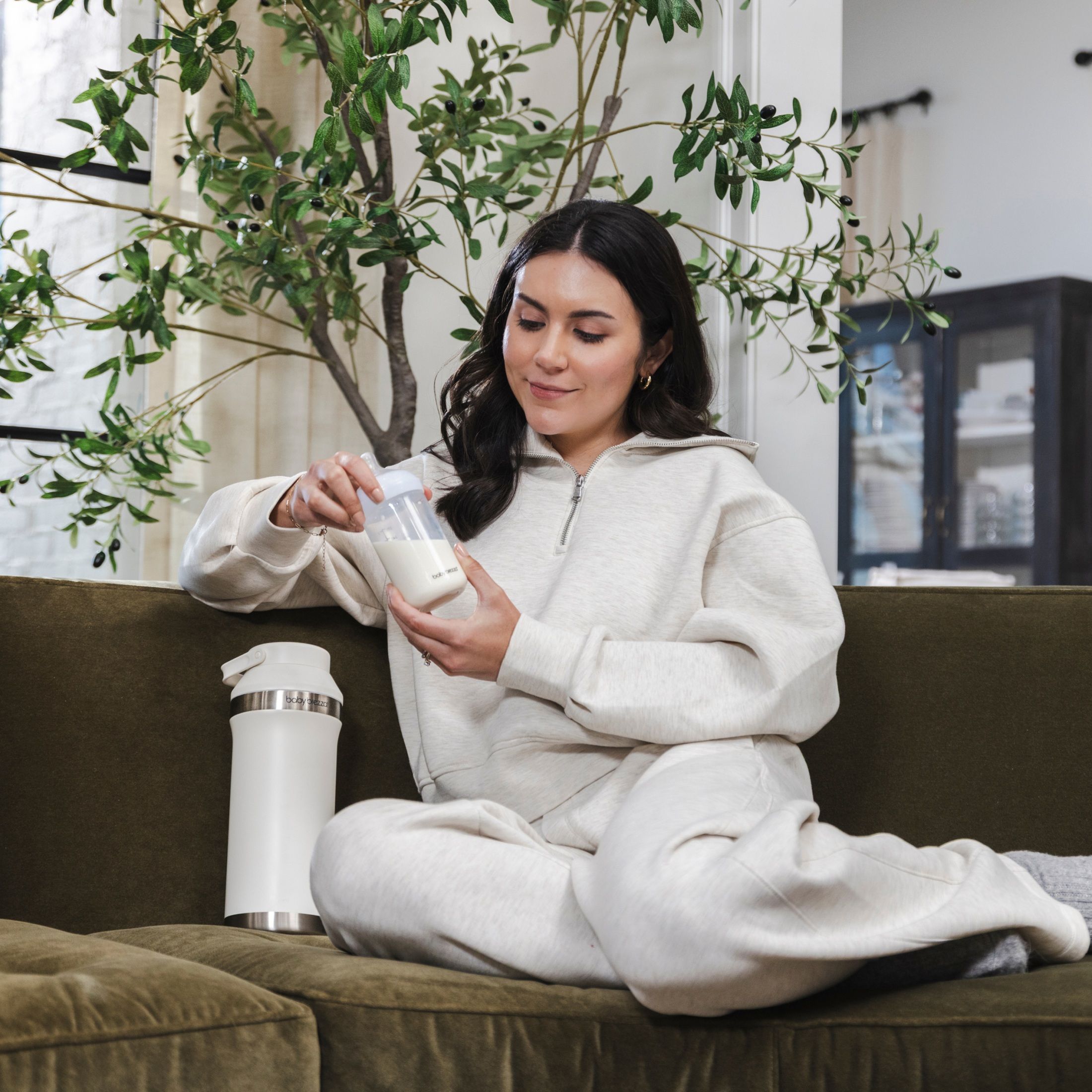 Eine Frau sitzt auf einem Sofa und hält eine Babyflasche mit Muttermilch in der Hand. Eine weiße Baby Brezza Thermosflasche steht daneben.