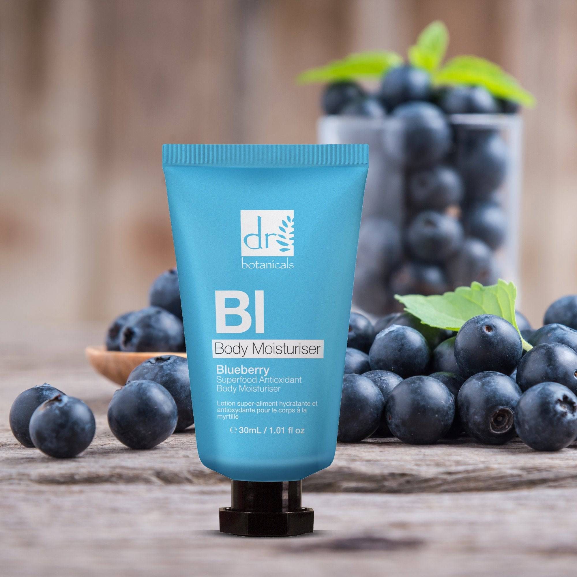 Blaue Tube vor Blaubeeren und Blättern. Text: Blueberry Superfood Antioxidant Body Moisturiser. Logo: Dr Botanicals. Auf Holzoberfläche.
