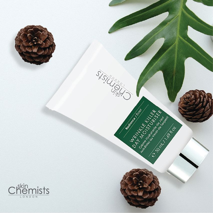 Weiße Tube mit grünem Etikett, umgeben von Tannenzapfen und Blättern. Aufschrift: Wrinkle Killer Day Moisturiser. Marke: skinChemists.