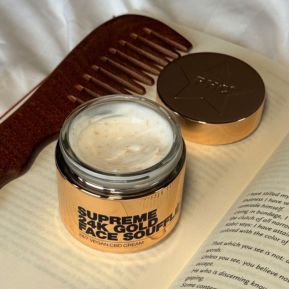 Goldfarbener Tiegel mit Creme, Deckel daneben. Aufschrift: SUPREME 24K GOLD FACE SOUFFLÉ. Offenes Buch und Kamm im Hintergrund.