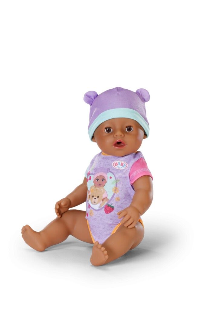 Puppe mit lila Mütze und Strampler. Die Puppe sitzt. Der Strampler hat ein Teddybär-Motiv.