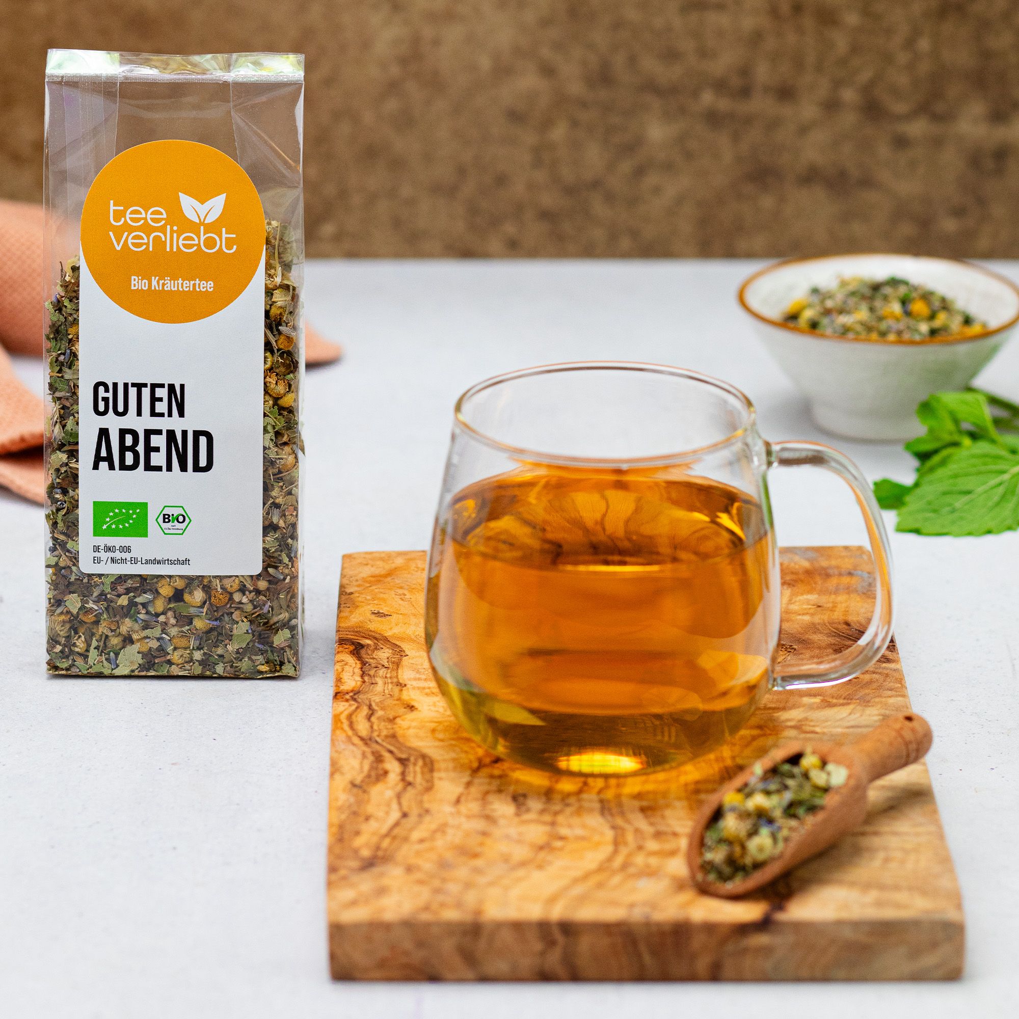 Verpackung mit Kräutermischung, Glasbecher mit Tee, Holzlöffel. Auf Holzbrett. Label mit Text 'Guten Abend'.