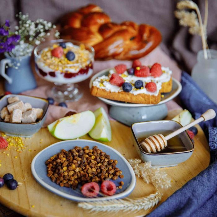 Holzbrett mit Speisen: Bienenbrot, Honig, Joghurt mit Beeren, Brot, Apfelstücke und Beeren.