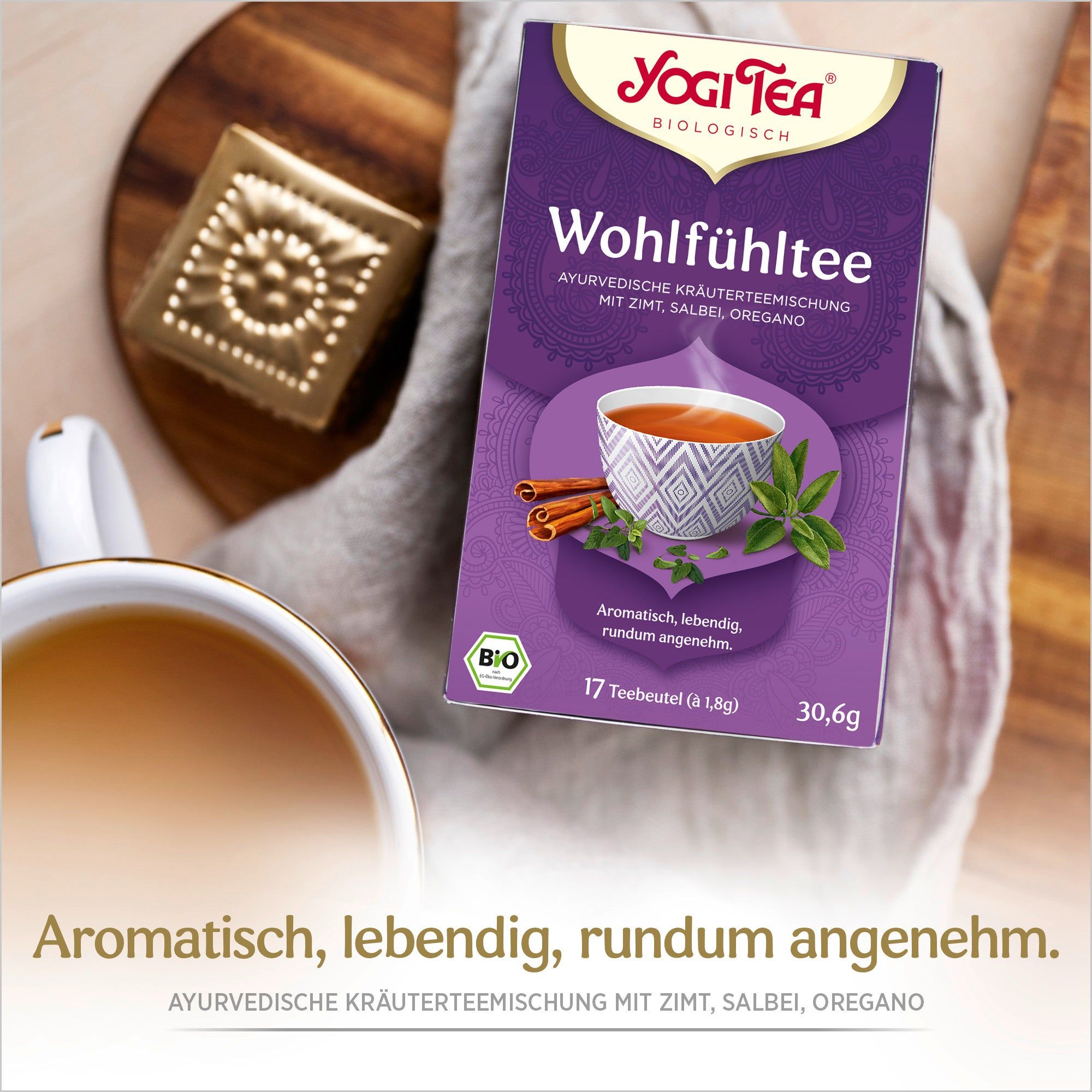 Yogi Tea Wohlfühltee-Packung auf Holzbrett. Lila Verpackung mit Logo, Produktname und Abbildung einer Tasse Tee mit Zimt und Kräutern. Bio-Siegel.