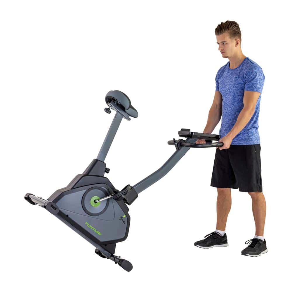 Mann hebt zusammengeklapptes Heimtrainer. Graues Fahrrad mit grünem Akzent. Tunturi-Logo sichtbar. Person trägt Sportkleidung.