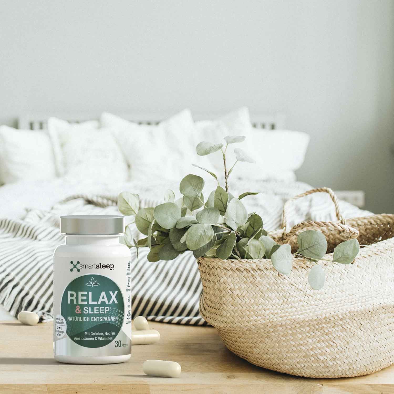 Weiße Flasche mit silbernem Deckel und Kapseln auf einem Tisch. Aufschrift: smartsleep RELAX & SLEEP. Dekoration mit Bett und Korb.