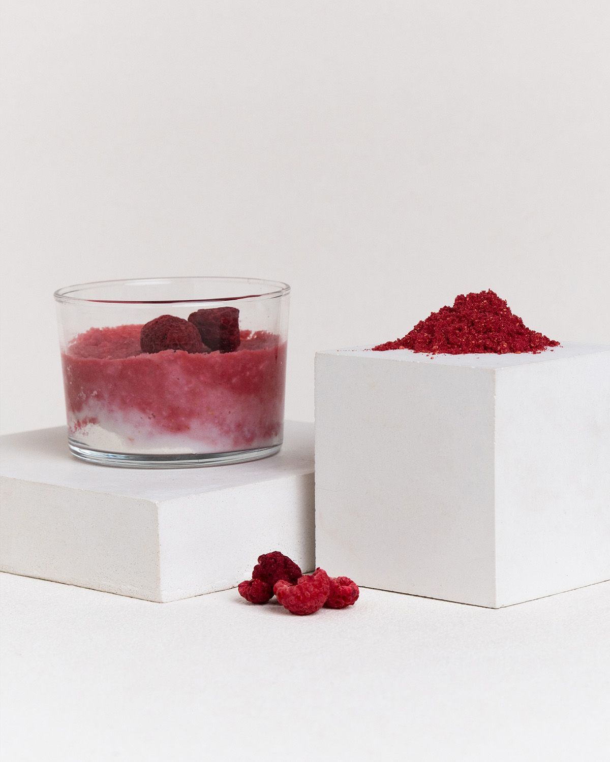 Himbeerpulver in einem Glas mit Joghurt und frischen Himbeeren, daneben ein Haufen Pulver.