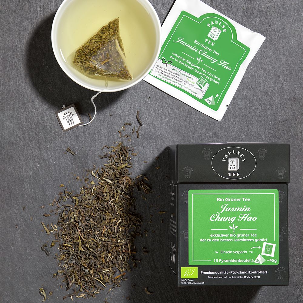 Tasse Tee mit Teebeutel und loser Tee. Teebeutel mit grünem Etikett. Aufschrift: Bio Grüner Tee Jasmin Chung Hao. Paulsen Tee Logo.