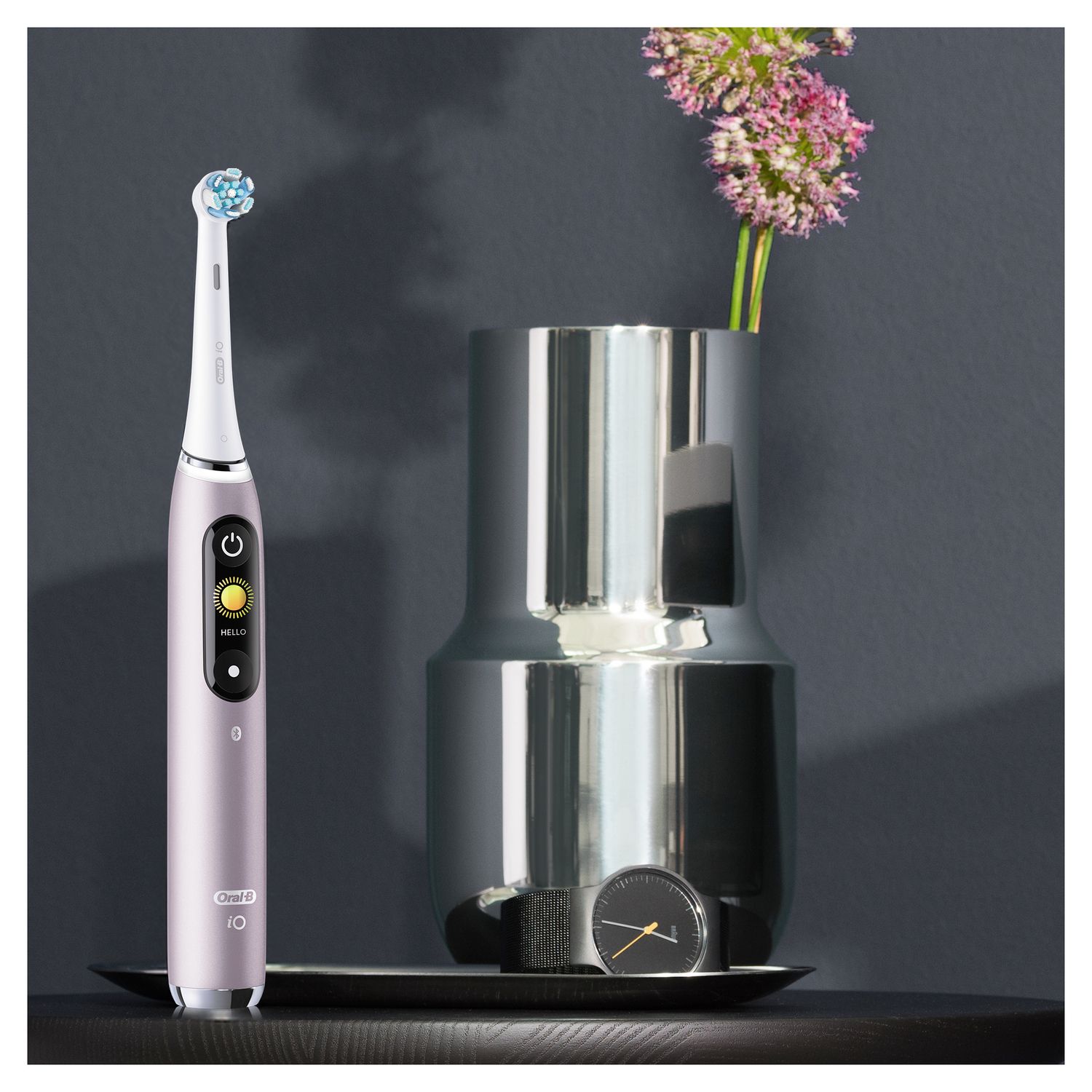 Elektrische Zahnbürste in Rose, neben einem silbernen Behälter und einer Vase mit Blumen. Schwarzer Untersetzer.