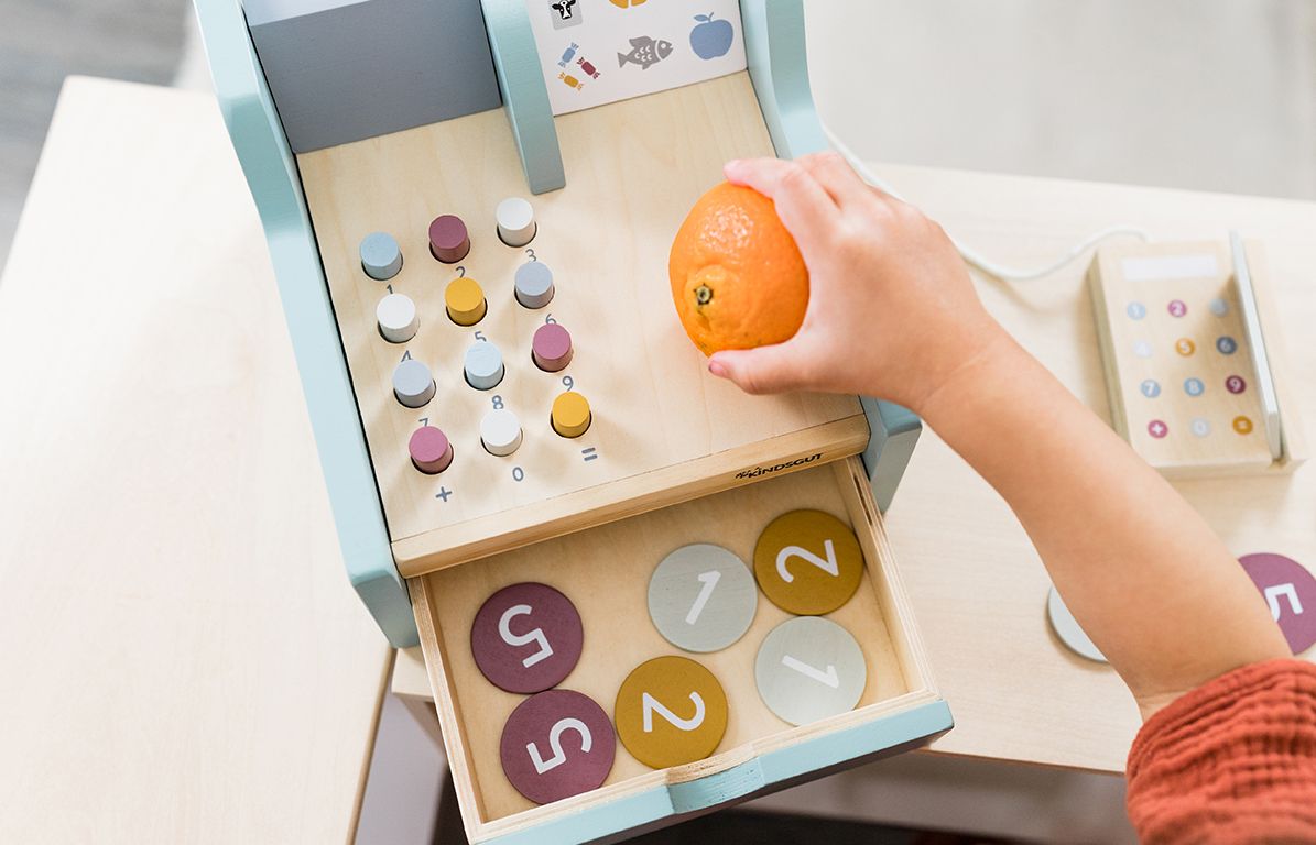 Kind hält Orange an Spielkasse. Kasse mit Knöpfen, Schublade mit Münzen. Scanner und Zubehör.