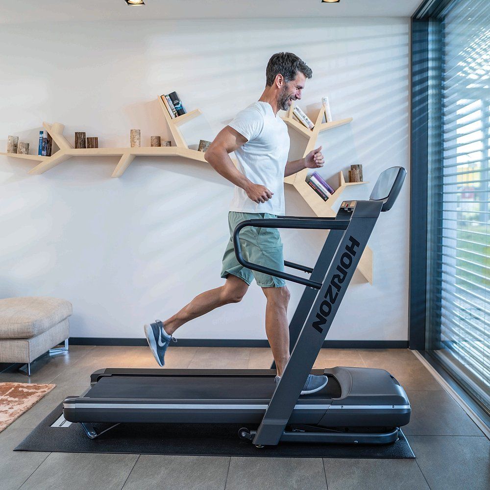 Mann läuft auf Laufband. Laufband: HORIZON Omega Z. Grauer Rahmen, schwarze Lauffläche. In Wohnraum.