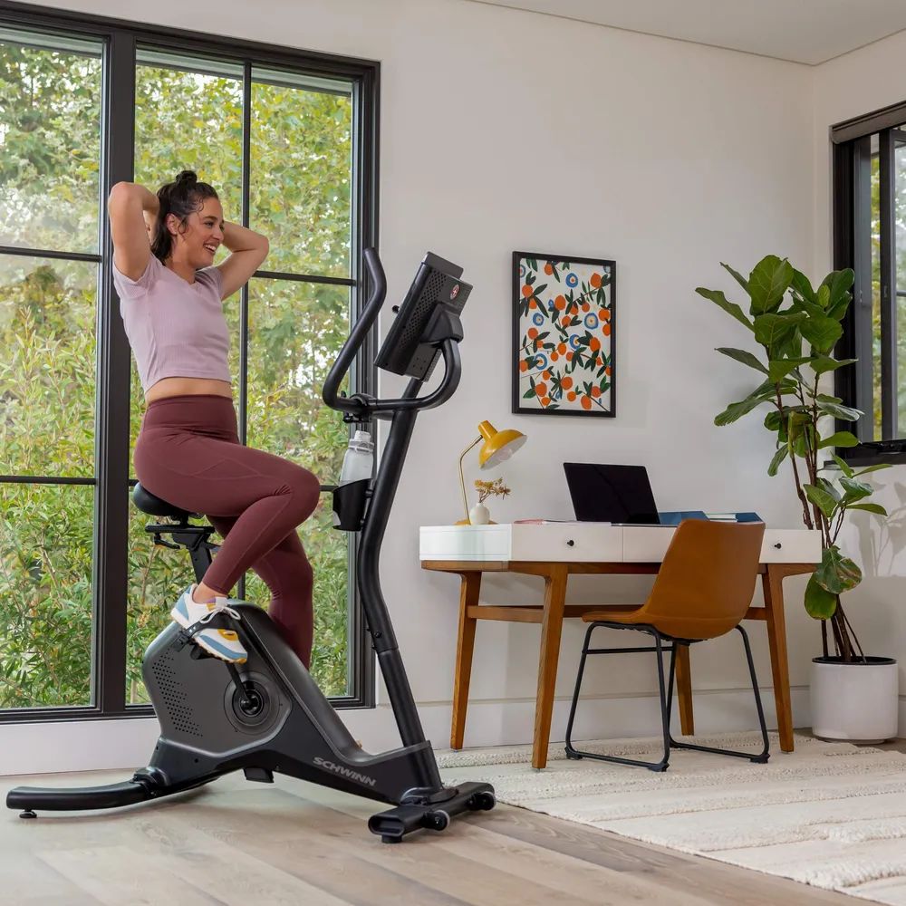 Frau trainiert auf einem Hometrainer. Schwinn-Logo. Innenraum mit Schreibtisch und Pflanze. Wasserflasche.