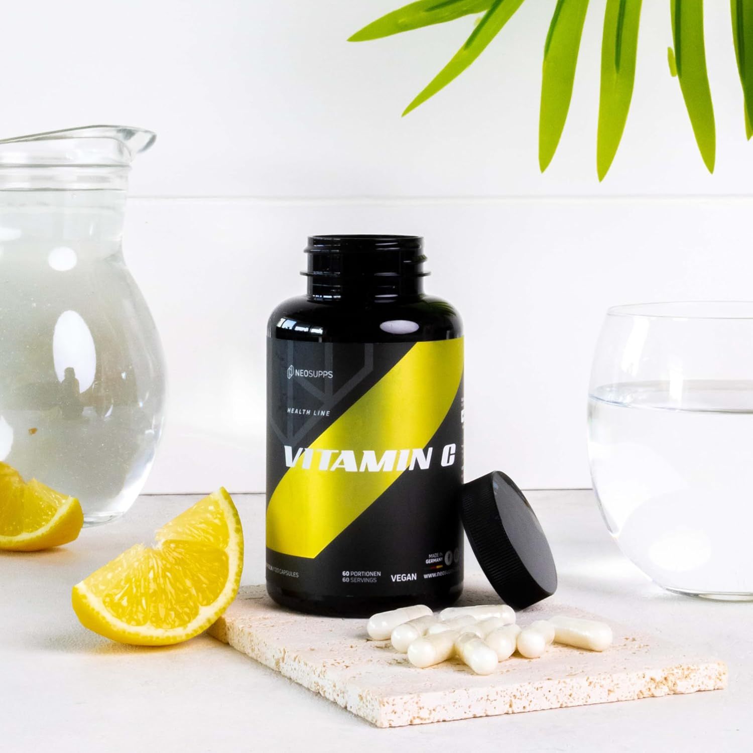 Schwarze Flasche mit gelbem Streifen und Aufschrift Vitamin C. Offener Deckel. Daneben Zitronenscheiben und Wasser.