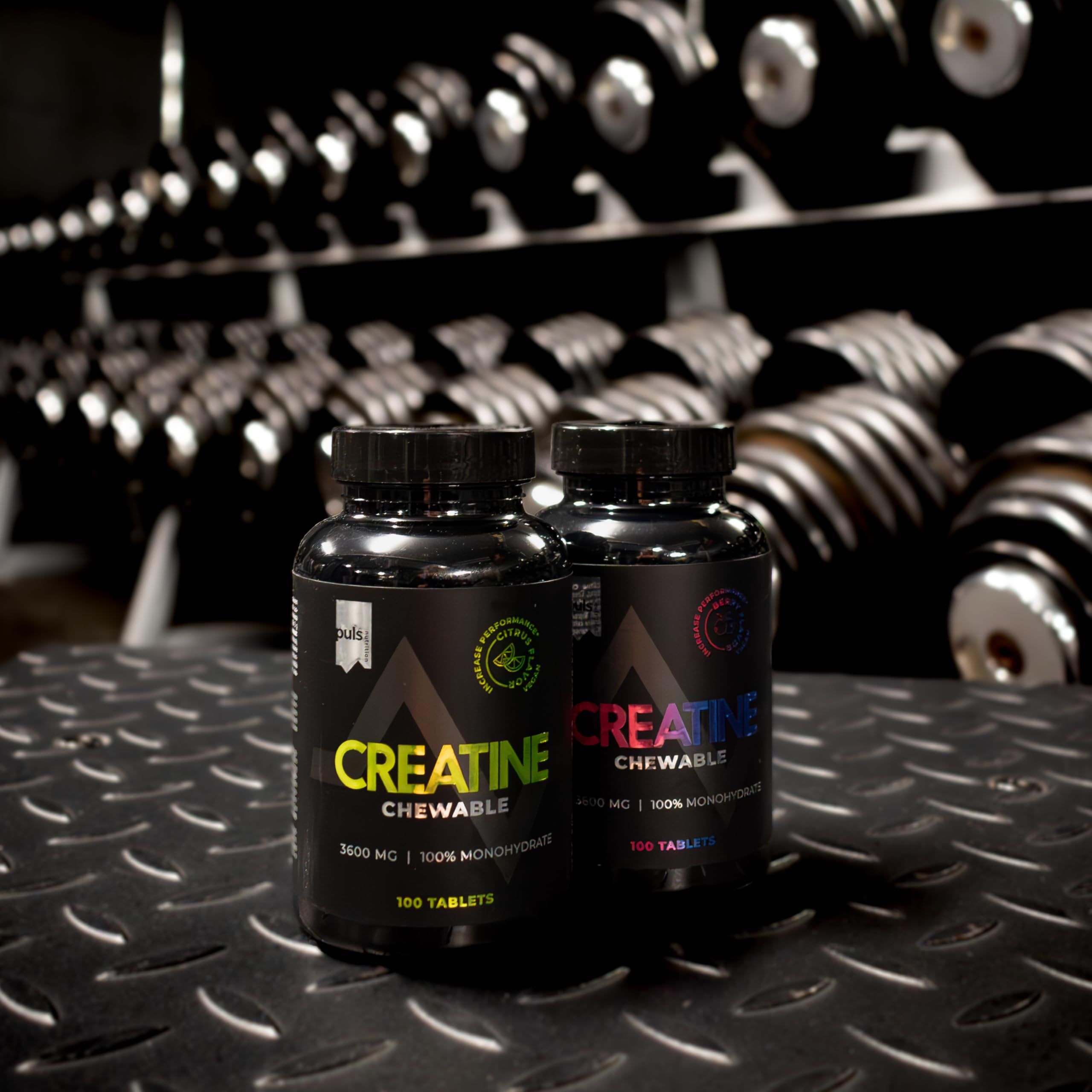 Zwei schwarze Dosen "Creatine Chewable" stehen vor einer Hantelablage im Fitnessstudio.