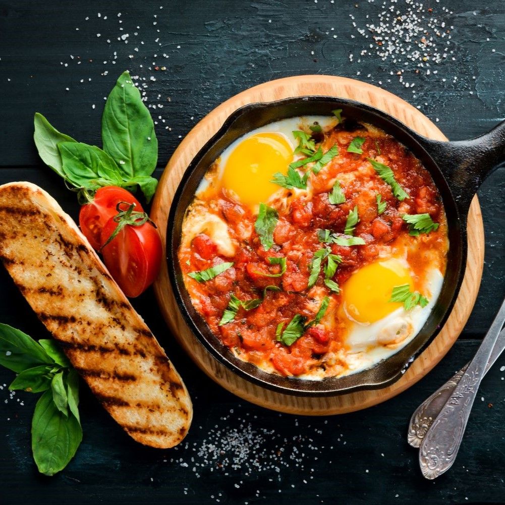 Gebratene Eier in Tomatensauce in einer gusseisernen Pfanne. Mit Brot, Tomaten und Basilikum dekoriert.