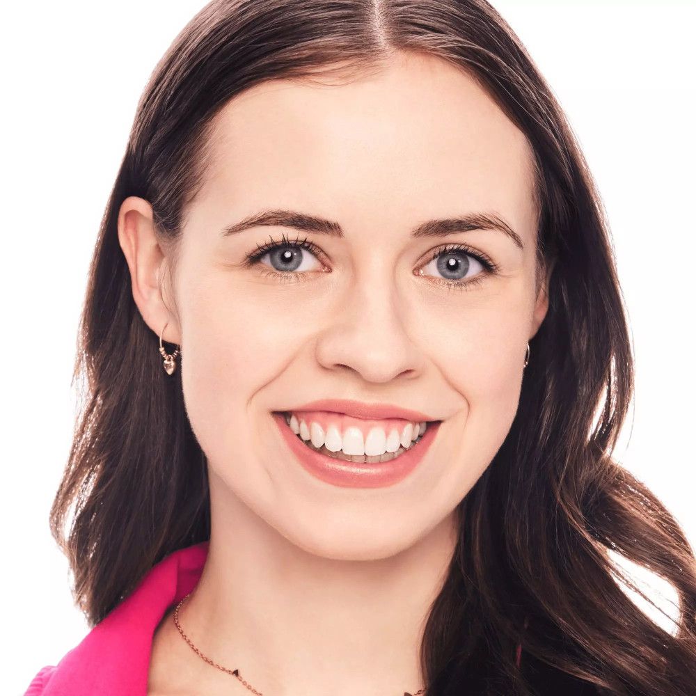 Porträt einer lächelnden Frau. Lange, braune Haare. Sie trägt einen rosa Blazer und eine Halskette.