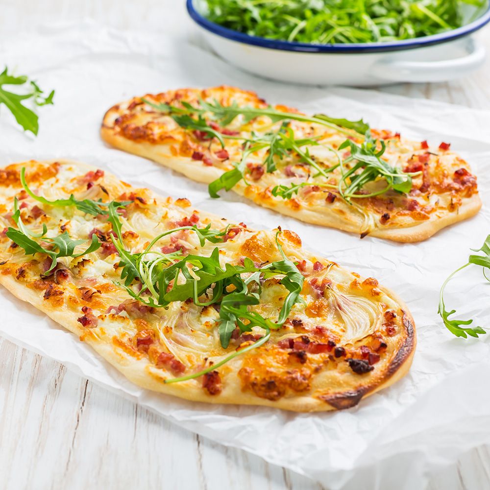 Zwei Flammkuchen auf Papier, mit Rucola belegt. Weiße Oberfläche, Schüssel mit Rucola im Hintergrund.