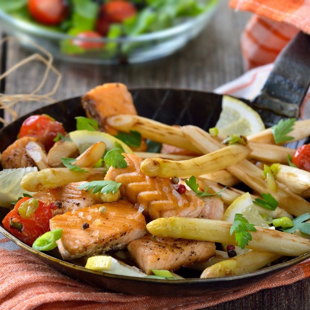 Gebratener Lachs mit Spargel, Tomaten und Zitrone in einer Pfanne. Auf einem Holztisch, mit einem orangefarbenen Tuch.