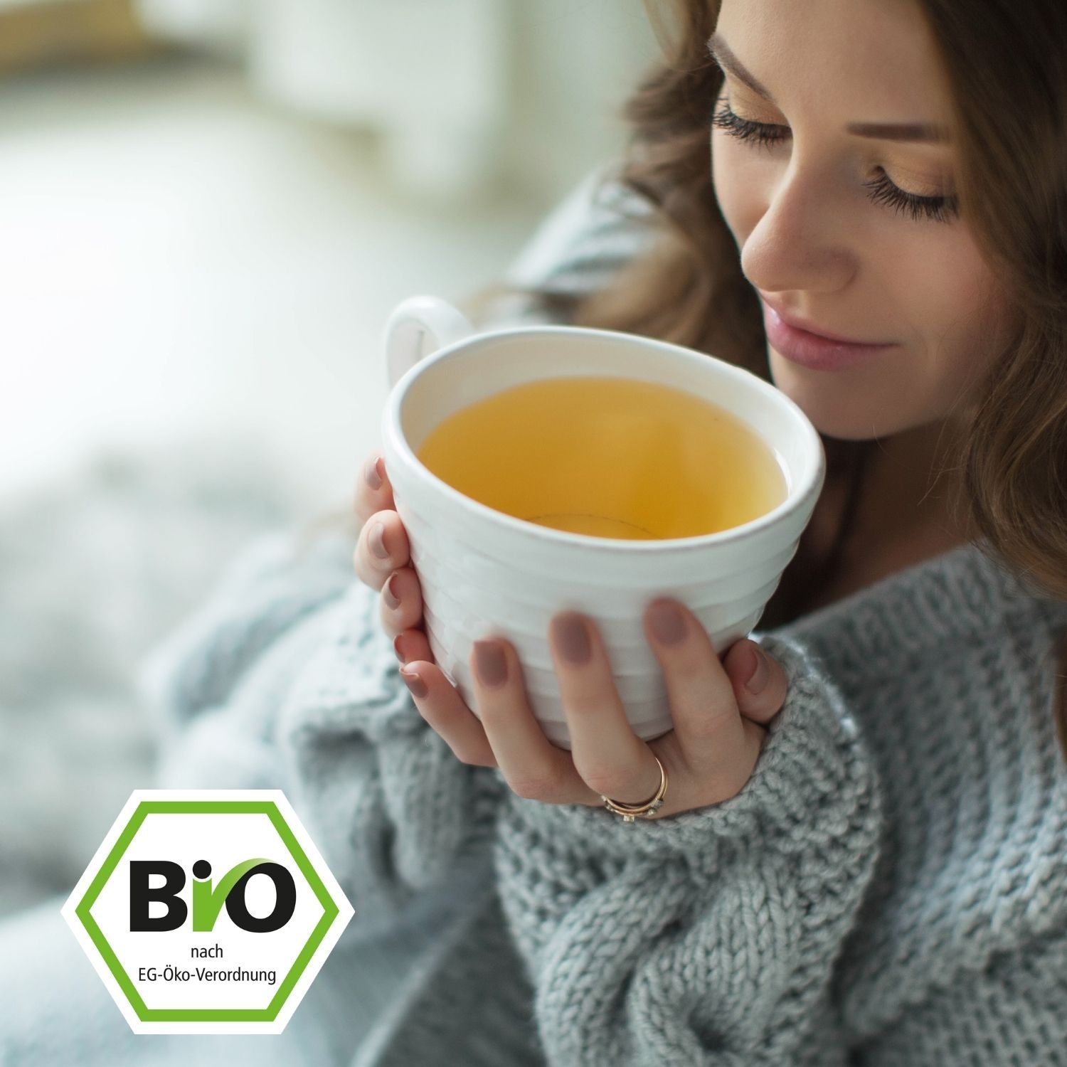 Frau hält eine weiße Tasse Tee. Bio-Logo im Vordergrund. Frau trägt einen grauen Pullover.