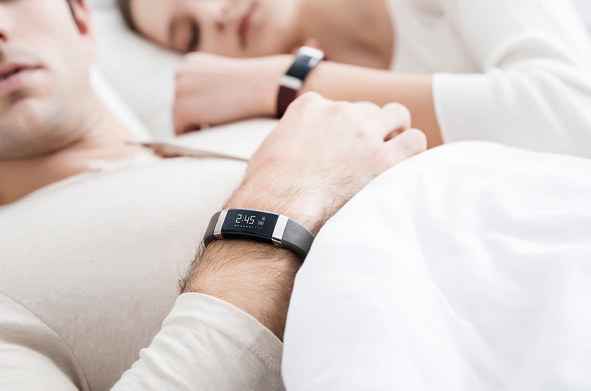 Personen im Bett mit Fitnesstrackern am Handgelenk. Schwarzes und braunes Armband. Zeigt Uhrzeit.