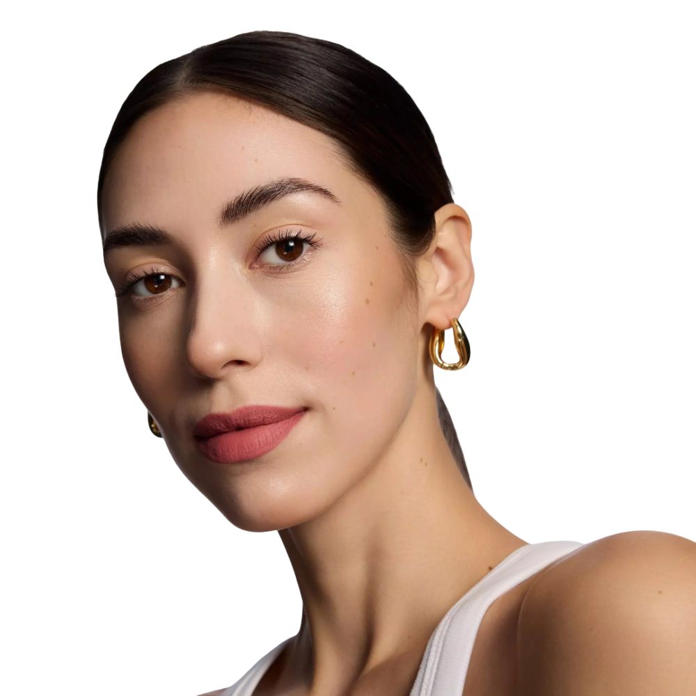 Porträt einer Frau mit rosafarbenem Lippenstift. Sie trägt goldene Ohrringe und ein weißes Oberteil.