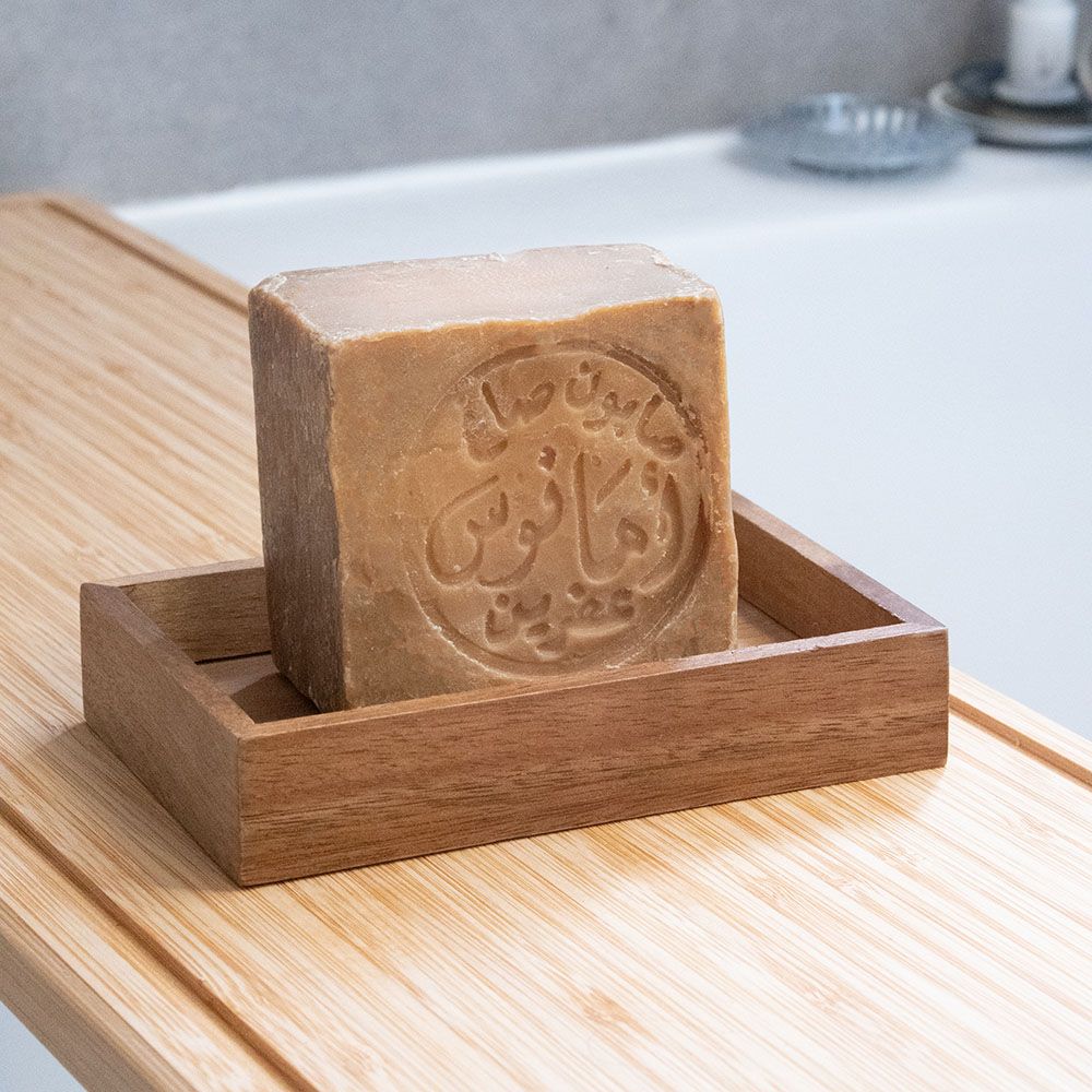 Een vierkant zeepblok met Arabisch schrift in een houten zeepbakje. Op houten plank.