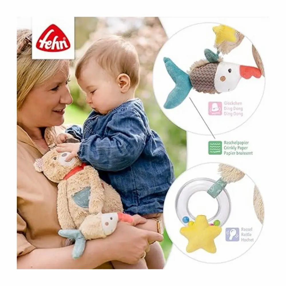 Baby hält Stofftier mit Fisch-Anhänger. Stofftier mit Mütze, rotem Schal, Stern. Marke: fehn. Mit Text.