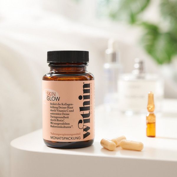 Braune Flasche Skin Glow, daneben Kapseln. Im Hintergrund weitere Flaschen. Auf weißem Tisch.