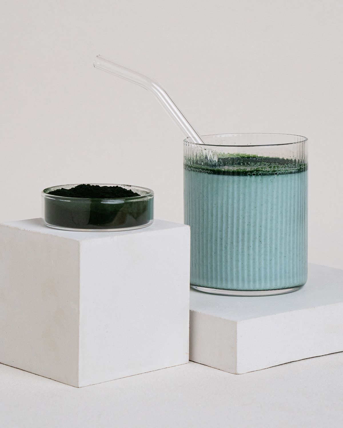 Ein Glas mit blauem Getränk und Spirulina-Pulver, daneben ein Schälchen mit Pulver. Ein Strohhalm steckt im Glas. Auf weißen Blöcken.