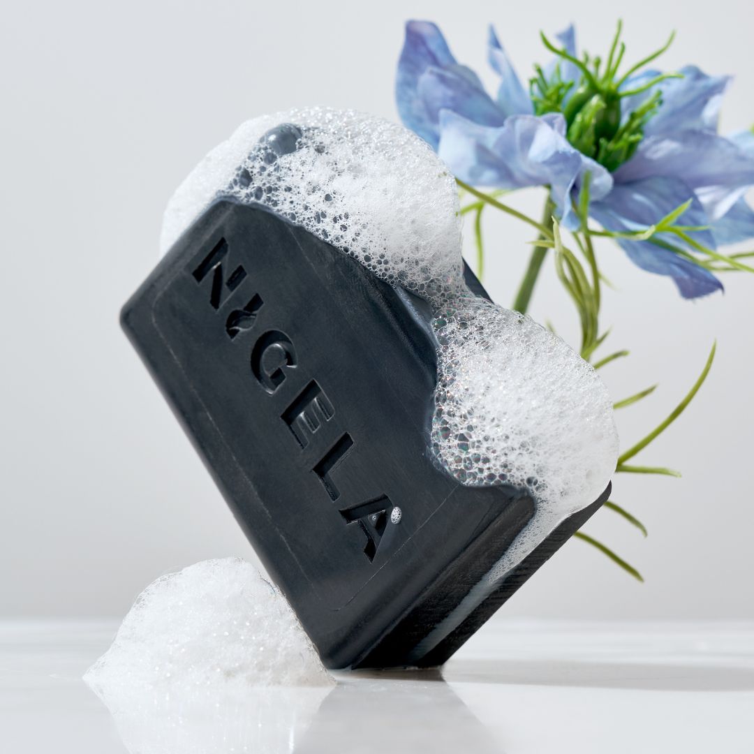 Schwarze Seife mit Nigela-Logo, mit Schaum bedeckt, neben blauen Blüten.