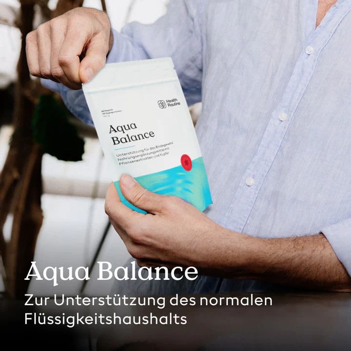 Person hält "Aqua Balance"-Beutel. Logo "Health Routine". Text: Unterstützung des Flüssigkeitshaushalts. Hintergrund: verschwommene Pflanzen.