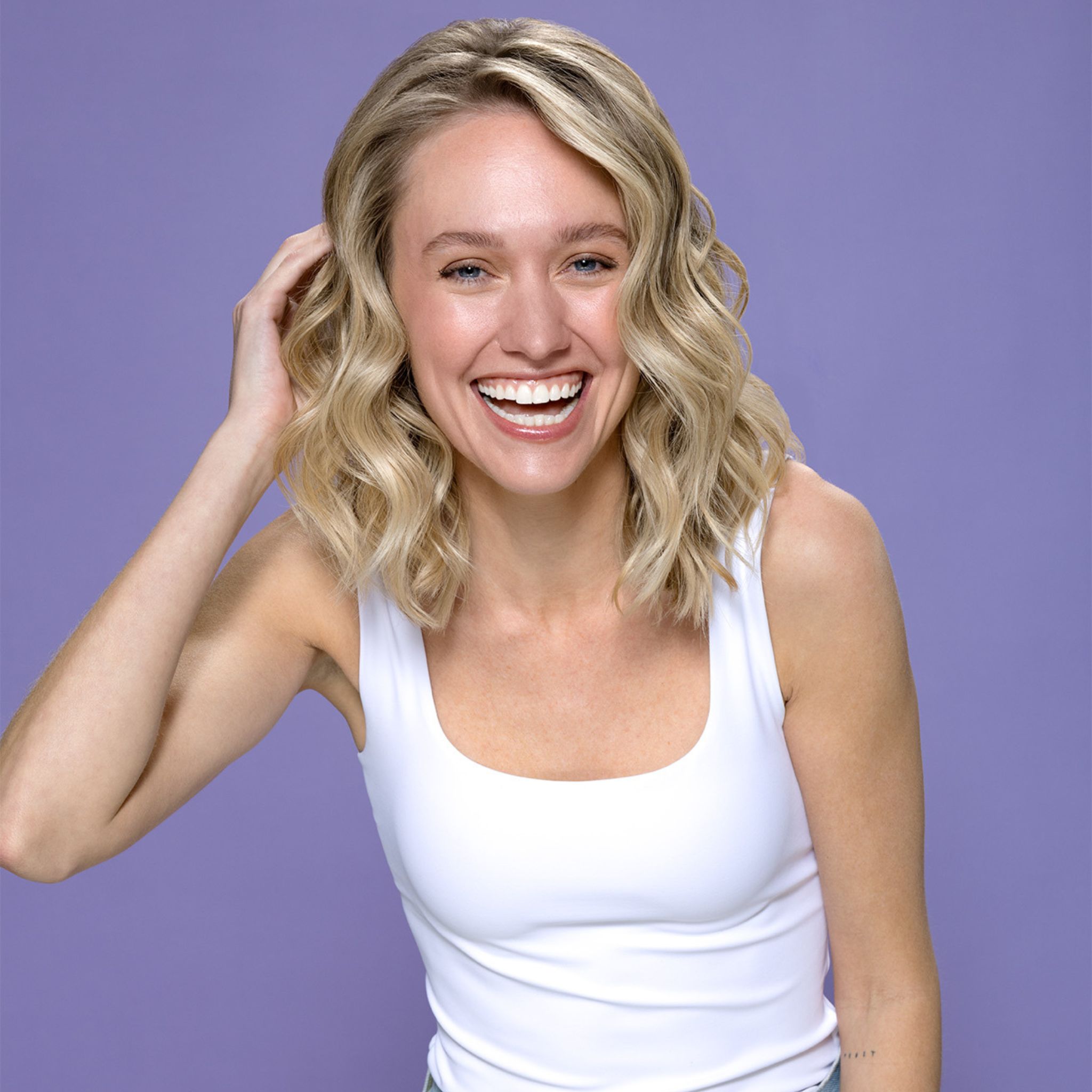 Eine lächelnde Frau mit blonden Haaren. Sie trägt ein weißes Tanktop. Der Hintergrund ist lila.