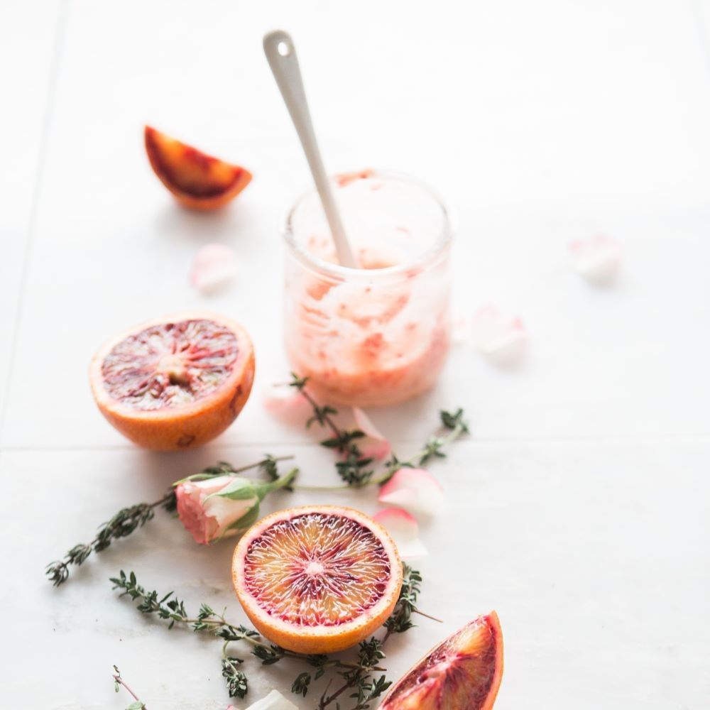 Halbierte Blutorange, Glas mit rosa Creme, Löffel, Thymian und Blütenblätter auf weißem Untergrund. Nahaufnahme.