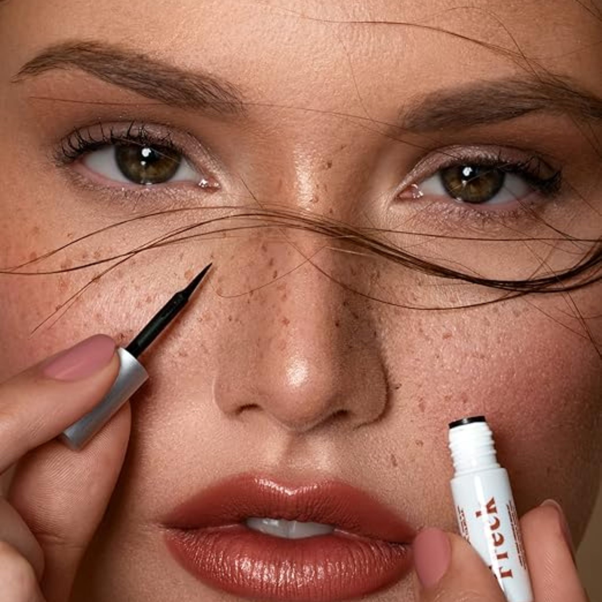 Primo piano di occhi e viso. Persona tiene eyeliner nero e tubetto con scritta "Freck".