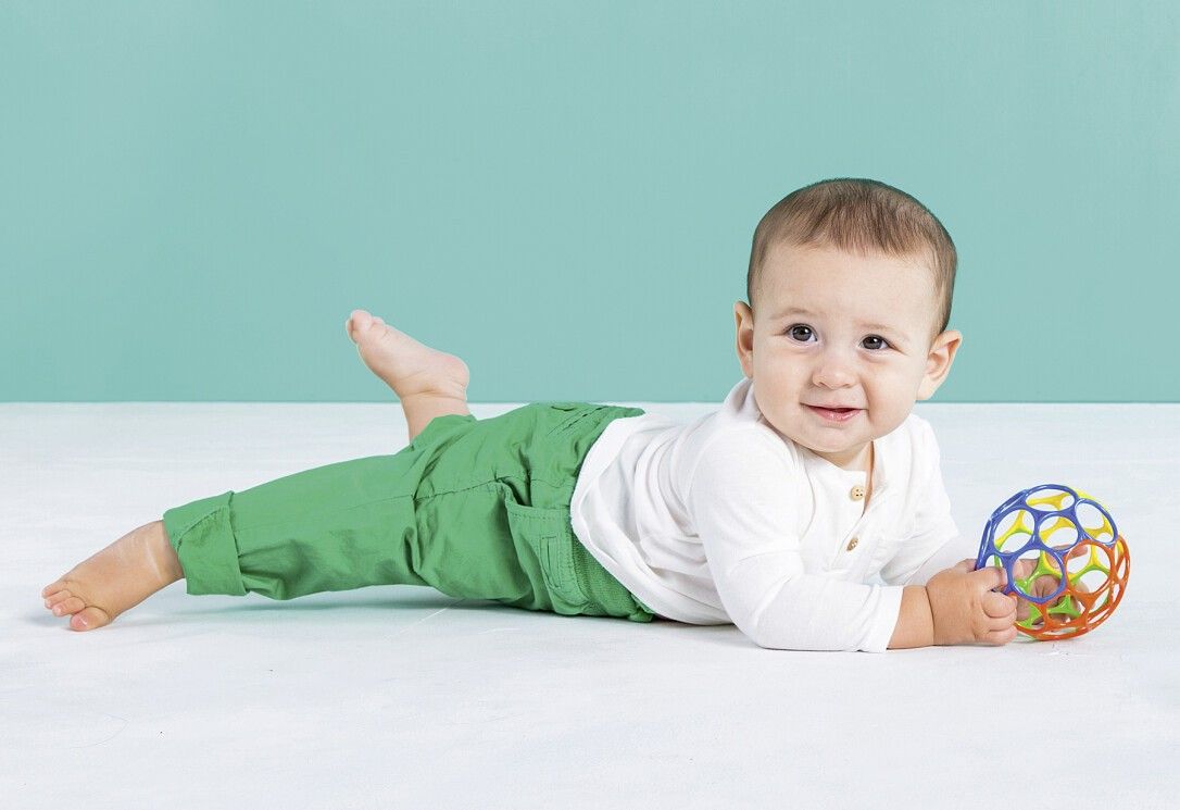 Baby liegt auf dem Bauch und hält einen mehrfarbigen Ball mit Löchern. Grün-blaue Umgebung.