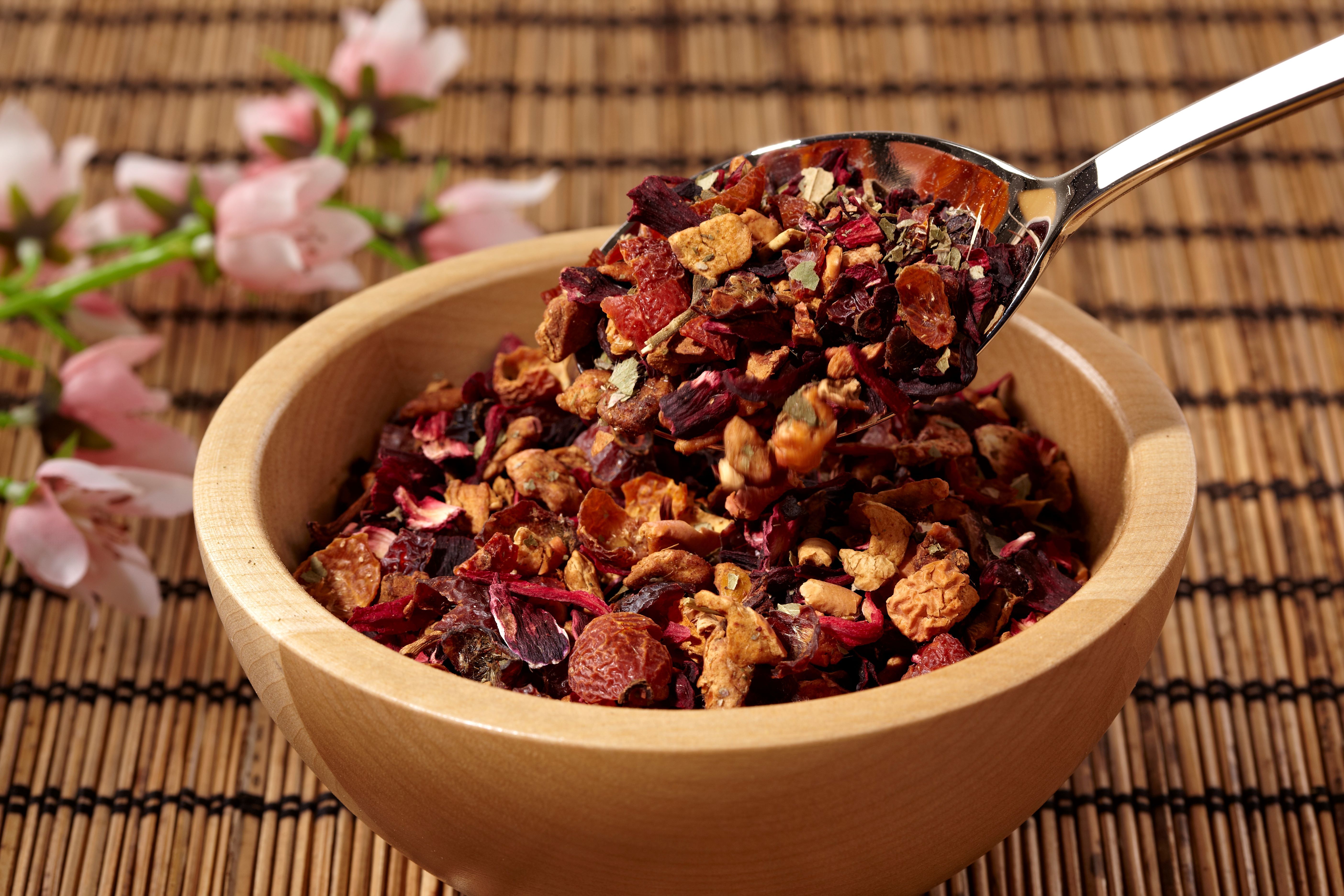 Holzschale mit Kräutermischung. Ein Löffel hebt Kräuter aus der Schale. Hintergrund: Bambusmatte und Blüten.
