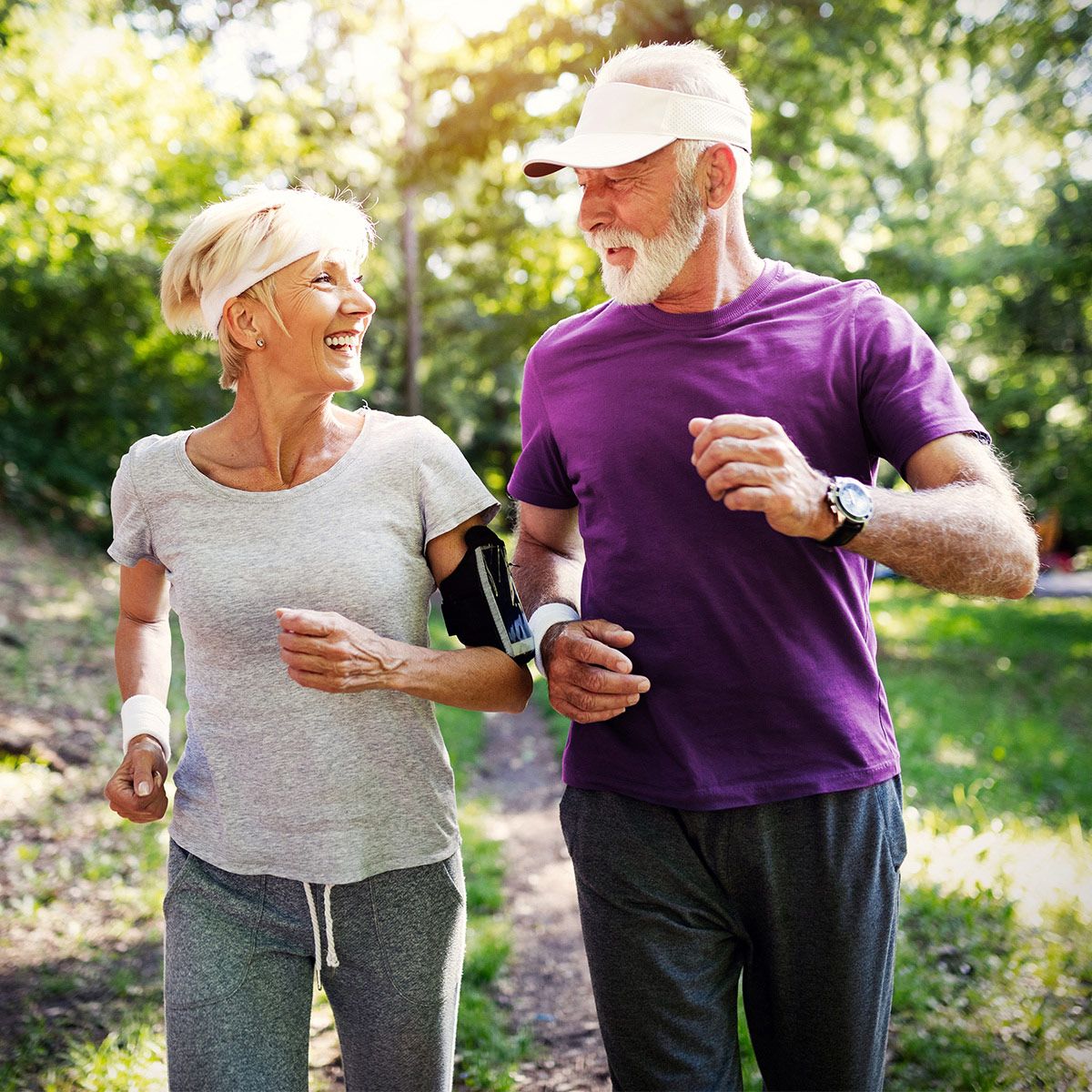 Älteres Paar beim Joggen im Park. Frau mit Stirnband und Armband, Mann mit Mütze und Uhr. Beide lächeln.